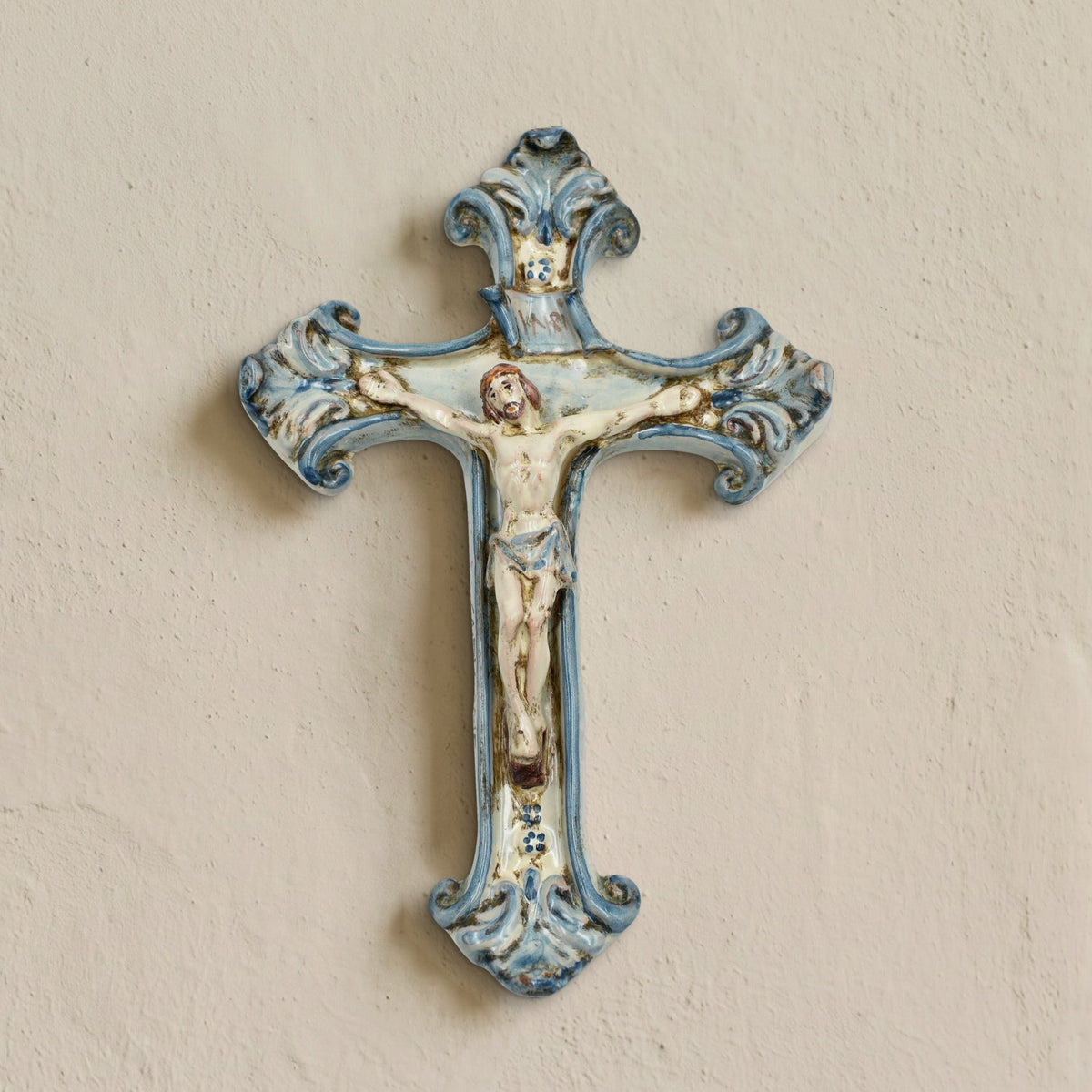 Crocifisso in ceramica di Caltagirone Mono Blu appeso a una parete chiara, fatto a mano con Cristo in rilievo e volute dipinte in blu
